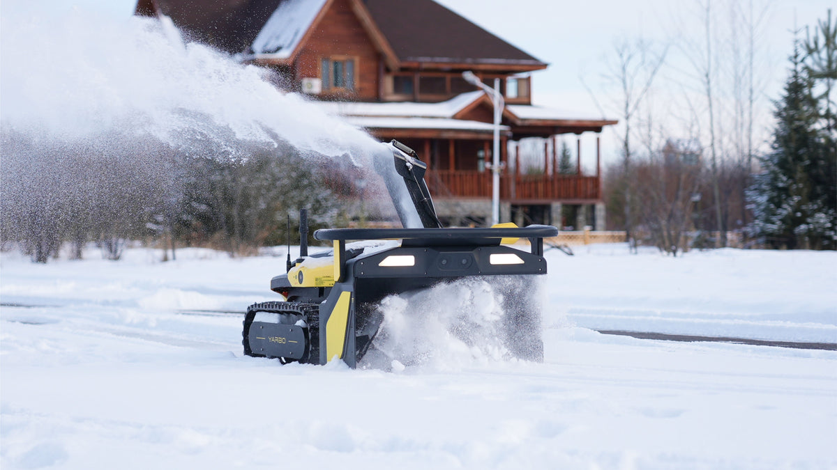 Refurbished yellow and black snow blower by Yarbo clearing snow in snowy yard, side view with illuminated headlights and tracked base